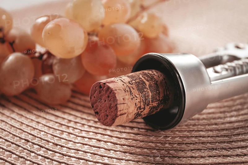A corkscrew with a wine bottle stopper, lying on a woven napkin, with green grapes