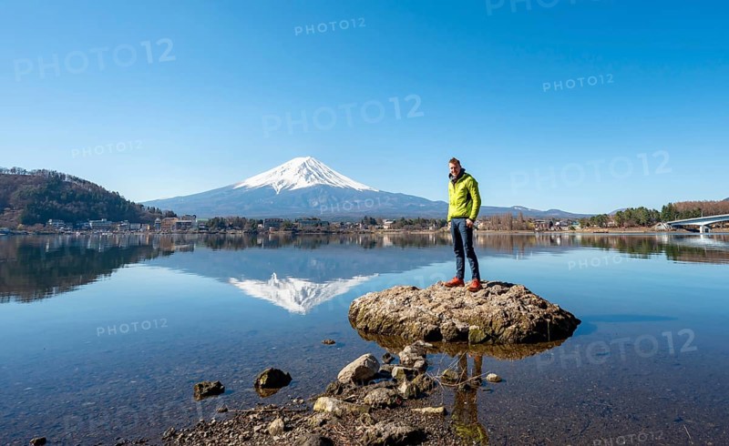 Young man standing on a rock in water, volcano Mt. Fuji is reflected in Lake Kawaguchi, Yamanashi Prefecture, Japan