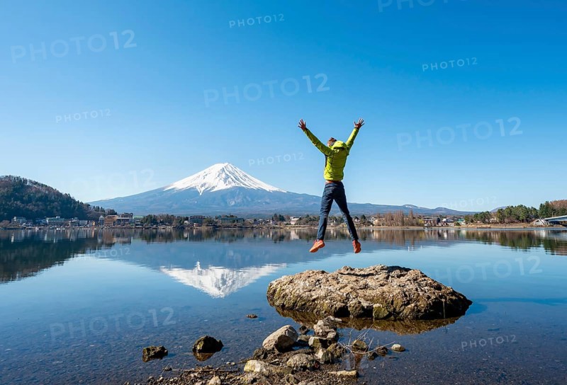 Young man standing on a rock in water, jumping into the air, volcano Mt. Fuji is reflected in Lake Kawaguchi, Yamanashi Prefecture, Japan
