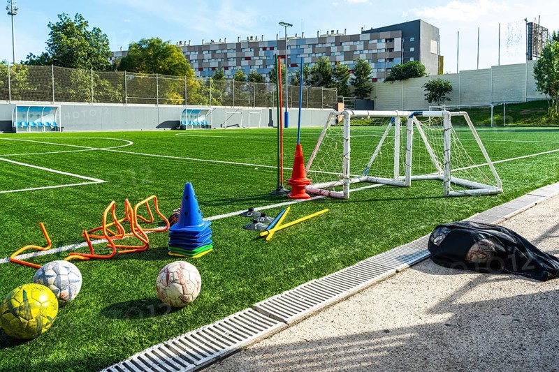 Artificial grass soccer field with multiple training equipment including cones, hurdles, a small goal, and soccer balls, ready for practice exercises under sunlight