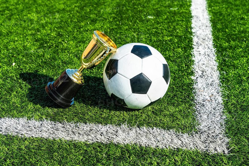 Soccer ball standing beside a gold champion trophy on a vibrant green artificial turf field with white lines, representing sports success, competition, and victory