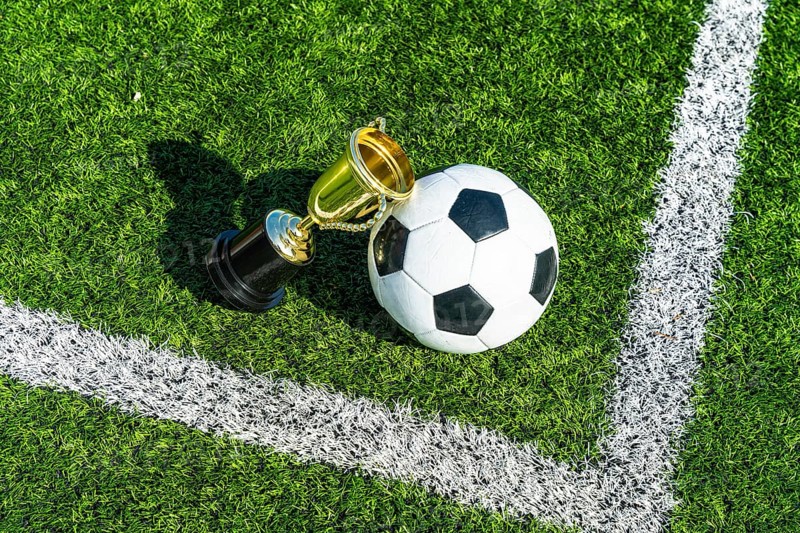 Soccer ball positioned alongside a gleaming gold trophy cup, symbolizing victory and championship success, resting on vibrant green artificial turf with white field marking