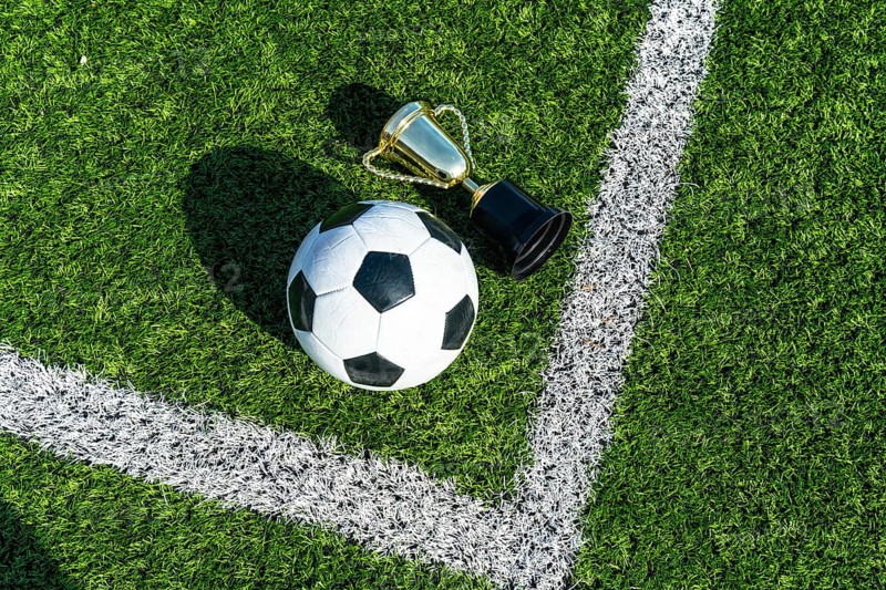 Soccer ball and gold trophy cup on artificial green turf with white line marking, top view flat lay evoking championship victory, success and sporting achievement on the pitch