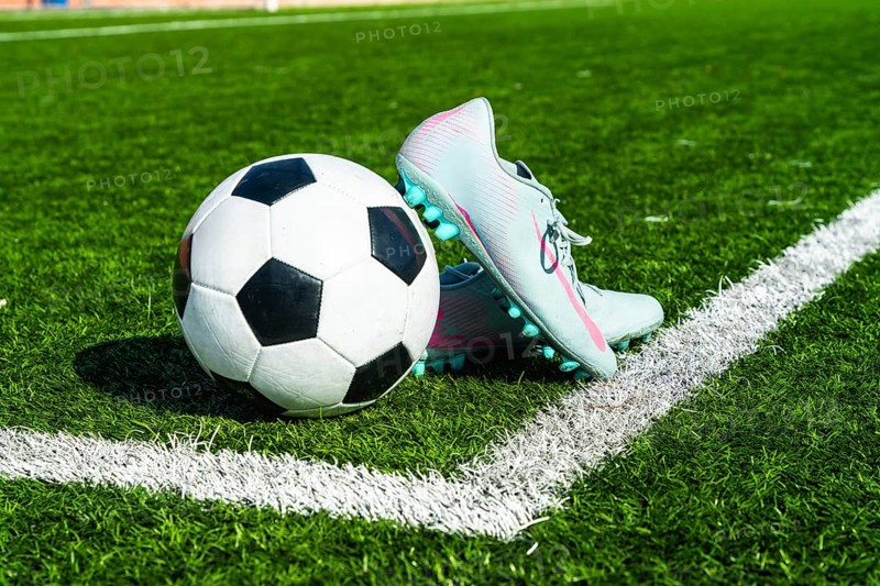 Soccer ball and football cleats are sitting on a green artificial grass pitch, close to the white boundary line of the vibrant playing field under sunny conditions