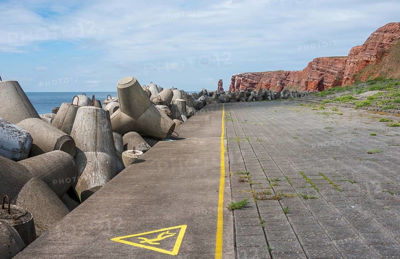 Coastline with concrete tetrapods by the sea, coastal protection, protective wall, paved coastal strip with yellow line, danger, warning, marking, red cliffs, cliff, cliffs, rock layers in the red sandstone and Lange Anna in the background, surf pillar, overcast sky, peaceful coastal scene, escape line, perspective, blue sea on the horizon, high seas island Heligoland, North Sea, Pinneberg District, Schleswig-Holstein, Germany