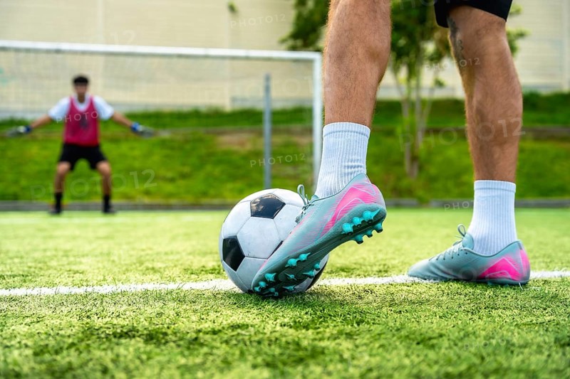 Soccer player's foot rests on a football, ready to take a penalty shot on a green artificial turf field, with a blurred goalkeeper standing in the background blocking the goal