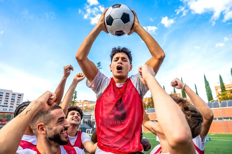 Young men soccer teammates in red bibs celebrate victory, raising a ball overhead with joyful cheers on a sunny stadium field, capturing teamwork and triumphant energy