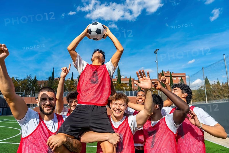 Young male soccer team members celebrating a goal or game victory on a green athletic field, lifting one player holding the ball high under a blue sky, all wearing red bibs