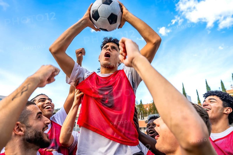 Young diverse men's soccer team members are cheering loud with fists raised and one player is triumphantly lifting a football overhead, symbolizing unity and achievement