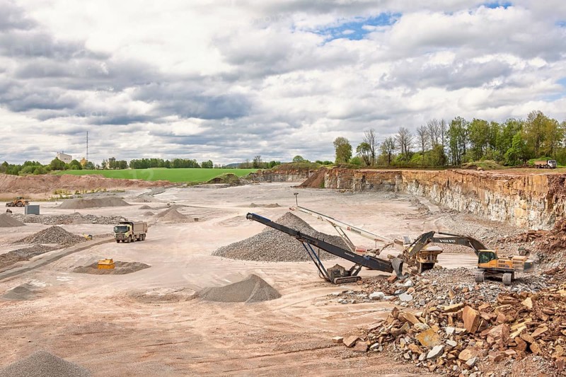 Excavator at a stone crusher with a conveyor in a limestone quarry with a waiting truck in a rural landscape, Sweden