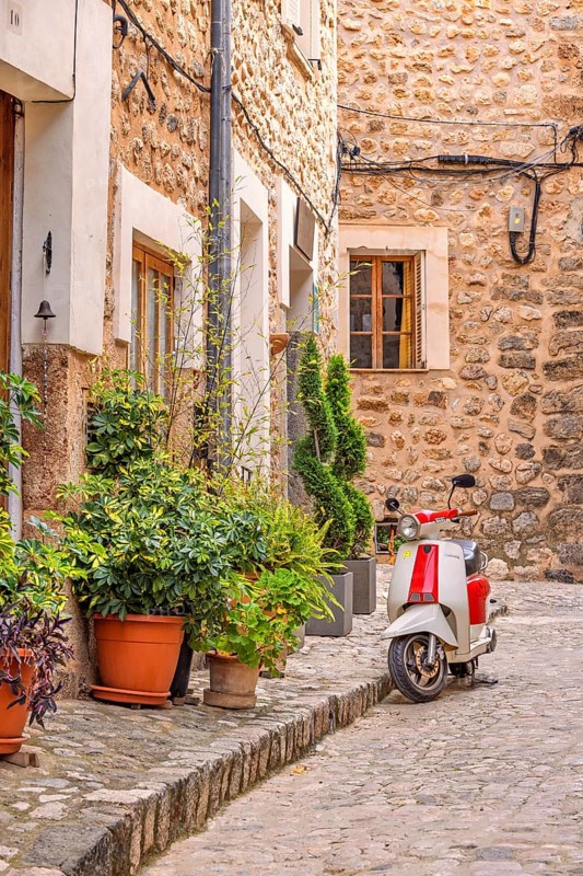Scooter parked on a street in an idyllic city street with green plants in front of the houses on the sidewalk, Soller, Mallorca, Spain