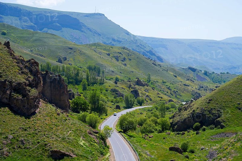 A winding road leads through a green valley with hills and trees under a blue sky, landscape between Aghitu and Vorotnavank Monastery, Vorotnavank, Syunik Province, Caucasus, Armenia