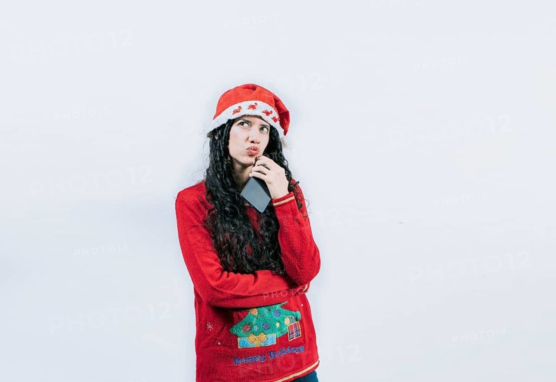 Young woman in christmas sweater thoughtful holding cell phone isolated. Girl in christmas hat thinking holding phone
