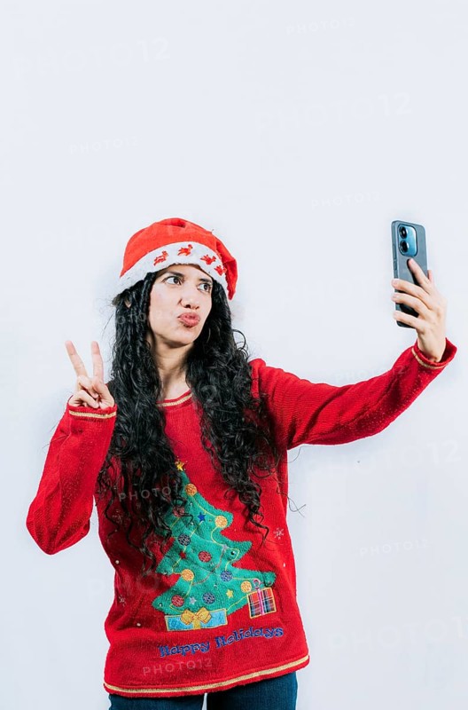 Funny latin girl in Christmas hat taking selfies and making gestures isolated. Cheerful young woman in Christmas sweater taking selfies