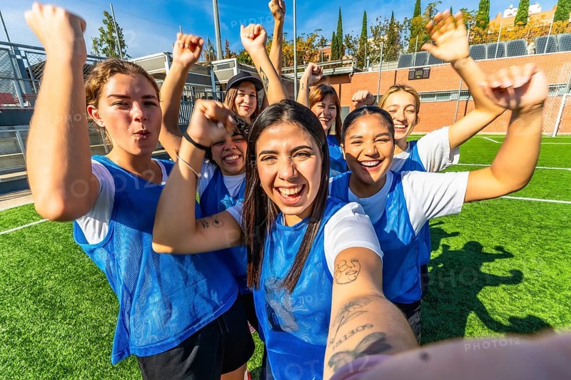 Happy young women's soccer team members celebrate a sports victory together on the green turf, raising their fists in excitement and taking a happy group selfie