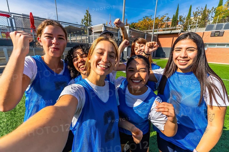 Group of smiling young women players celebrating success and victory on a soccer field, posing for a selfie after a match, showcasing teamwork and friendship