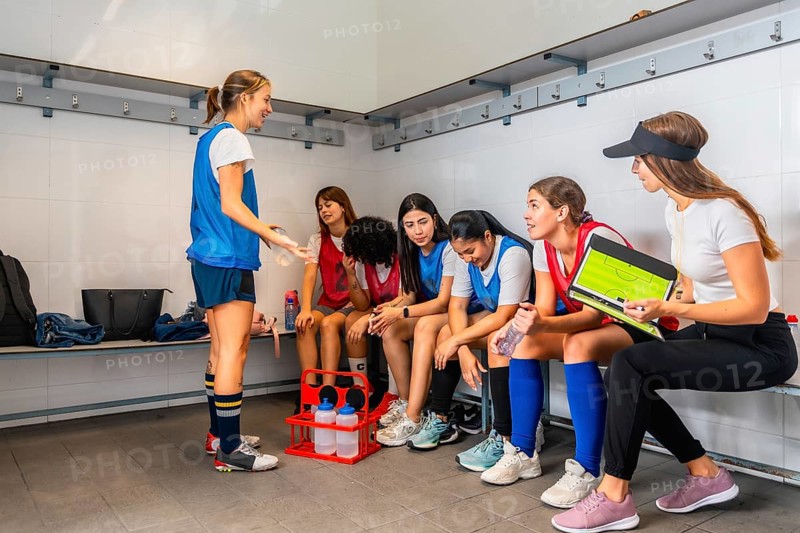 Coach explaining game strategy on a clipboard to a diverse group of young women soccer players listening attentively in the locker room, emphasizing teamwork and communication