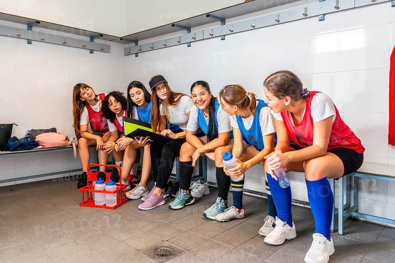 Female athletes wearing sports attire sitting on a bench in a locker room, discussing strategy and reviewing a folder before or after a sports event, showing teamwork and communication