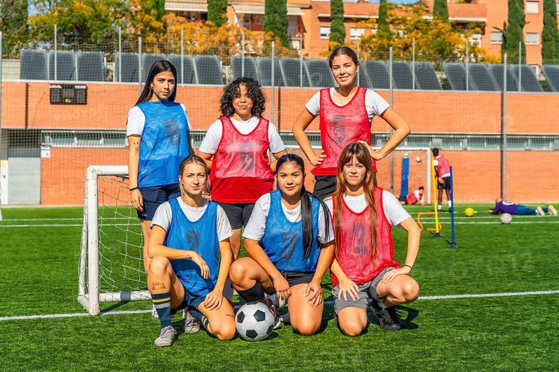 Diverse group of women on a soccer field, some standing and some crouching, wearing jerseys and bibs, confidently posing as a team before a match or training session