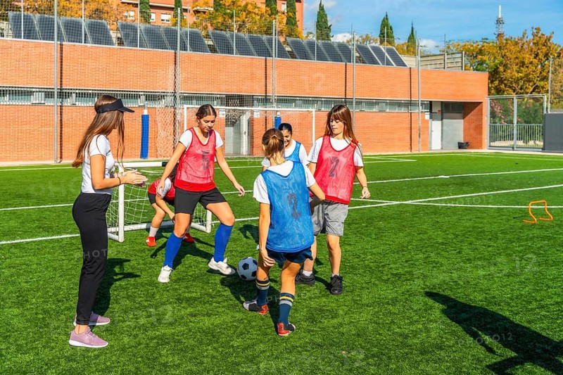 Girls soccer team receiving instructions from their female coach while practicing ball control and passing drills on an artificial turf field, developing teamwork and football skills