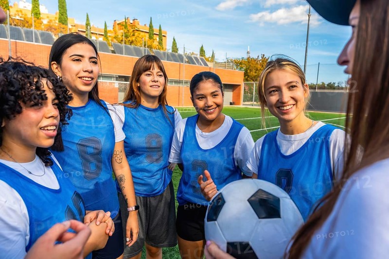 Diverse women football players smiling and listening, holding a soccer ball on a green artificial turf field, preparing for a match or training session