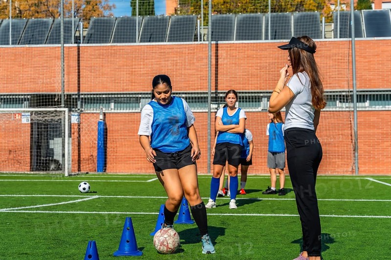 Female soccer players receiving instructions from their coach during a practice session on a green turf field, focusing on individual ball control drills