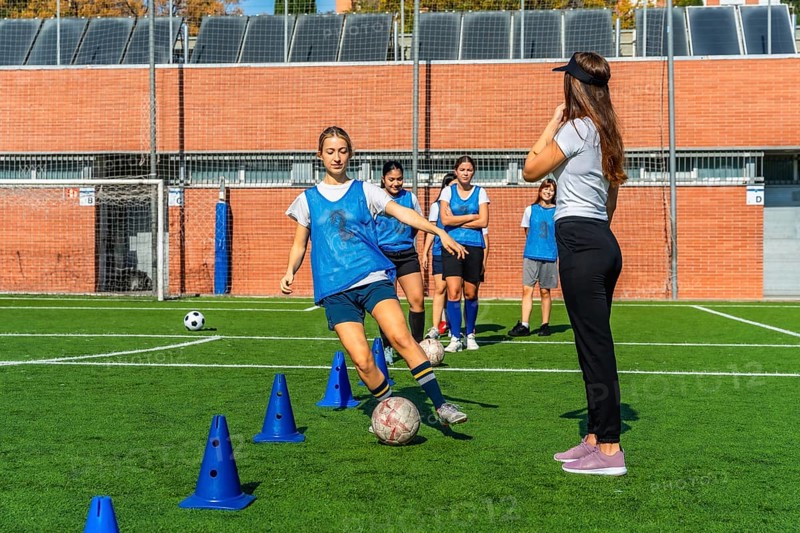 Group of diverse young women soccer players training with their coach on a green artificial turf field, practicing dribbling skills around blue cones during a sports session