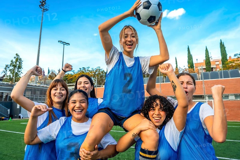 Group of diverse women soccer players cheering and lifting a teammate with a soccer ball on a green field, expressing happiness and teamwork after a competitive match