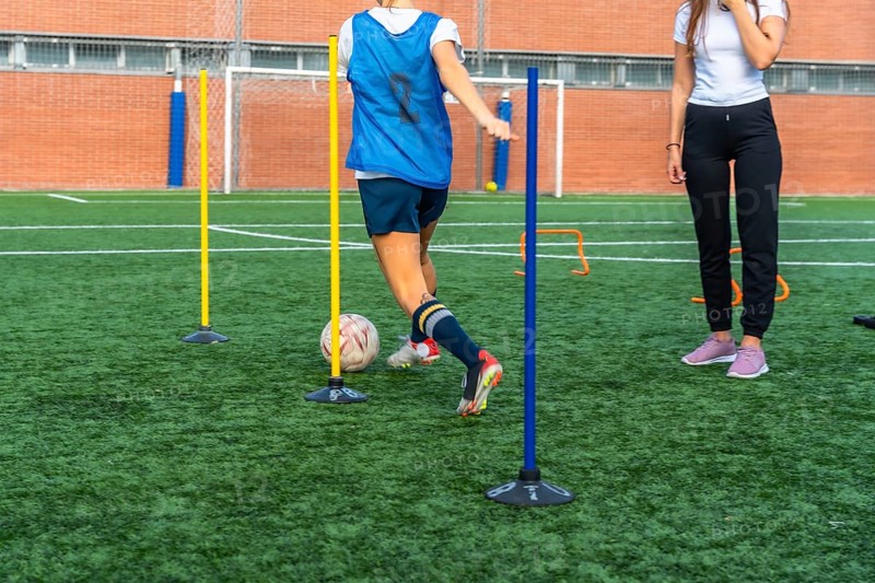 Young woman in a blue bib practicing soccer dribbling skills, maneuvering a ball through training poles on a vibrant green artificial turf field with a coach observing
