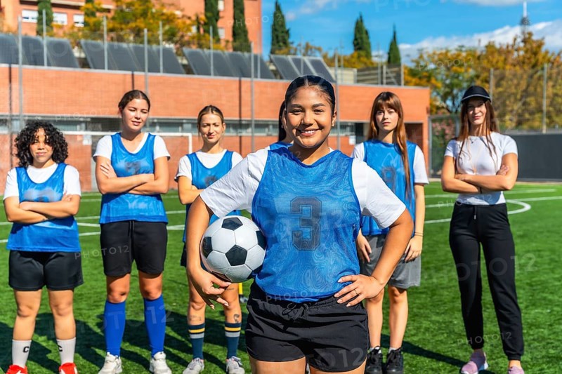 Diverse women's soccer team, featuring a smiling player holding a football, standing proudly on a green sports field, representing teamwork, sport, and female empowerment