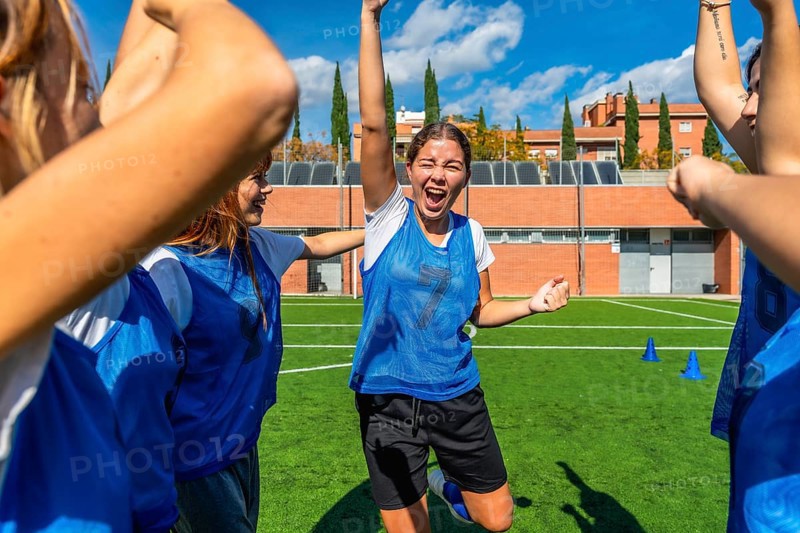 Young women's soccer team celebrating a goal, arms raised and cheering with bright smiles on a sunny field, showing teamwork, victory and joyful energy