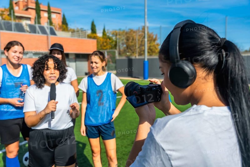 Videographer with headphones filming a reporter holding a microphone and interviewing a group of diverse young women soccer players on a green sports field