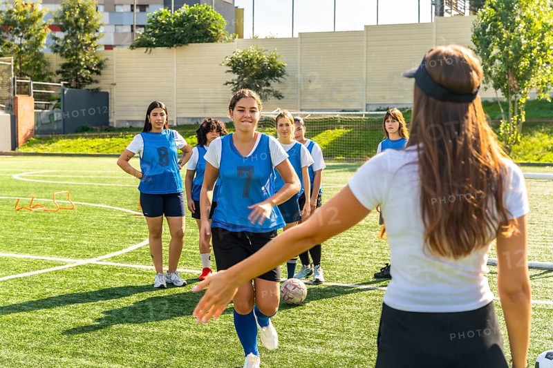 Young women's soccer team participating in a training session on a sunny artificial turf field, receiving instructions from their female coach while practicing their skills and teamwork