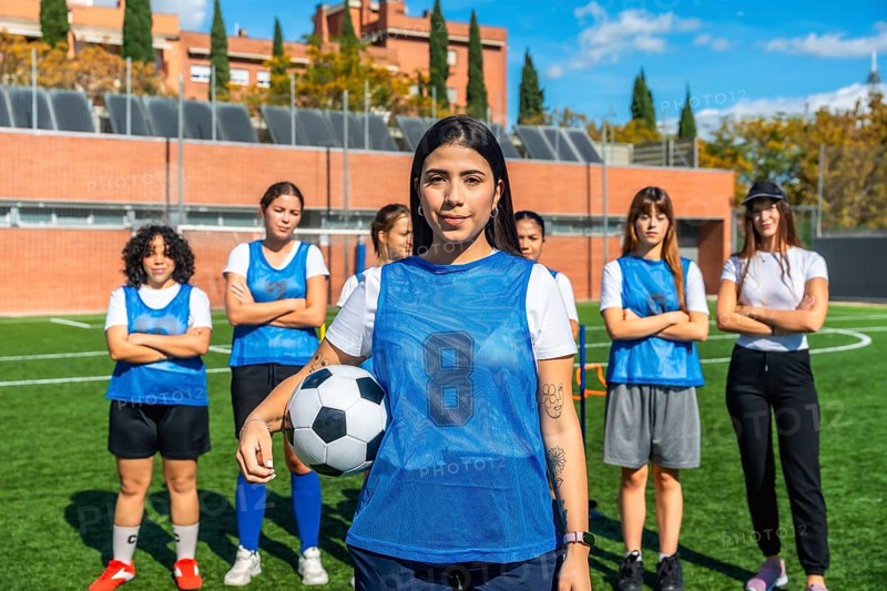 Young female soccer players standing on a sunny artificial turf field, with one player holding a football and looking at the camera, representing teamwork and active lifestyle