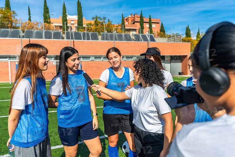 Women soccer team players standing on a green sports field, one player holding a microphone while interviewing two teammates, with a videographer filming the scene