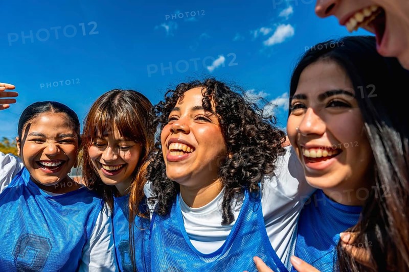 Diverse group of young girls standing close, smiling and laughing under a bright blue sky, embodying team spirit and fostering strong bonds of friendship and camaraderie after a sports game