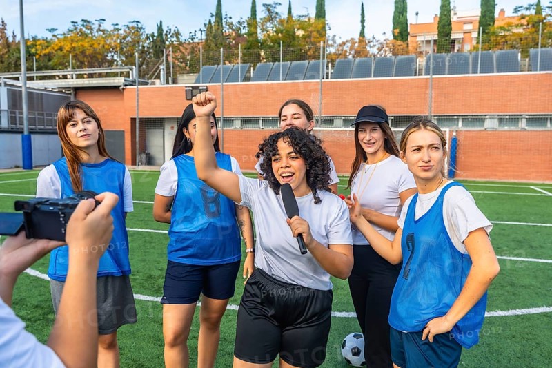 Happy young women's soccer team and coach giving an interview on artificial turf field, celebrating success and empowerment during a group conversation