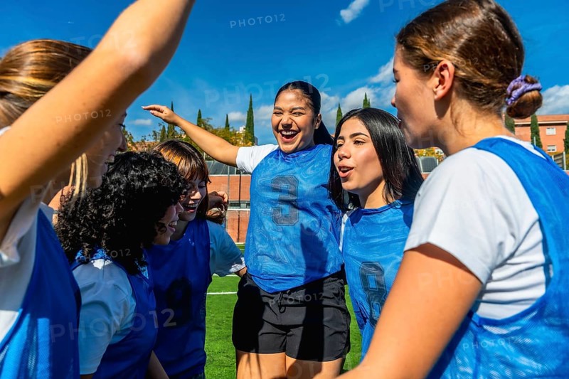 Group of diverse women's soccer players huddling on the field, actively celebrating their success and showing vibrant team spirit and strong teamwork under a bright blue sky
