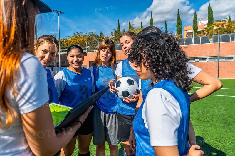 Women's soccer team huddling with their coach on a green artificial turf field, listening intently to stratagems and instructions during a training session