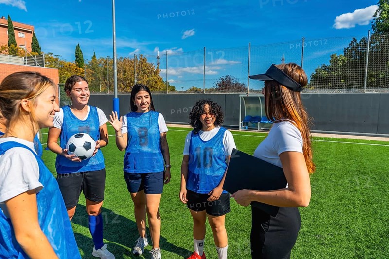 Women soccer players in blue pinnies listening attentively to their female coach explaining strategies and guiding the team on a sunny artificial grass field