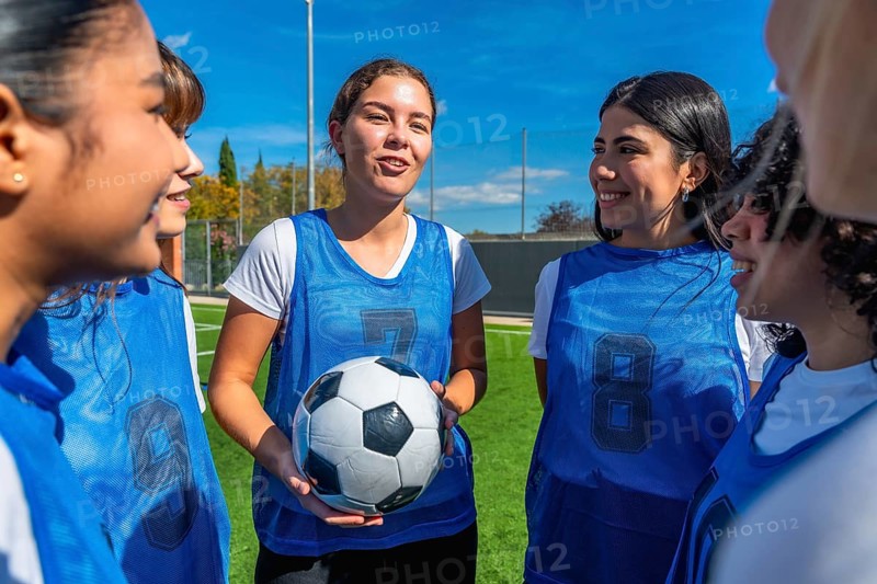 Young women soccer players huddling on a green field under a clear blue sky, one player holding a soccer ball while actively listening and smiling with her teammates