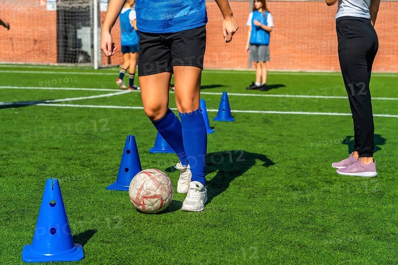 Young girl in a blue jersey practicing soccer dribbling skills around orange cones on a green artificial turf field, training for the sport with her team on a sunny day