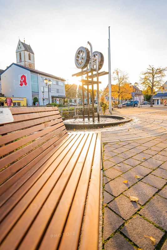 Municipal square with church and sculpture at sunset, concrete paving and autumn trees, Aidlingen, Böblingen district, Germany