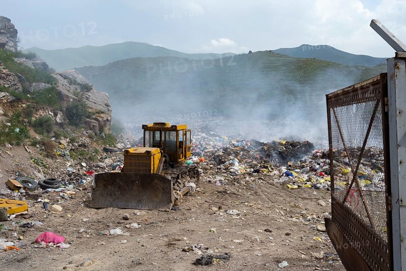 An excavator on a smoking garbage dump in the middle of mountains under a cloudy sky, Syunik province, Armenia