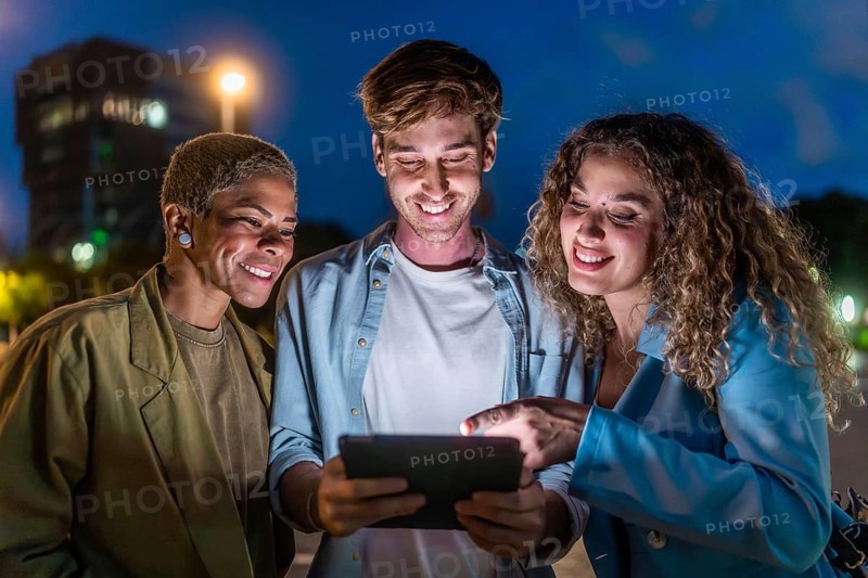 Three smiling diverse friends standing outdoors at night, looking at a shared digital tablet screen while interacting and enjoying content together in an urban setting