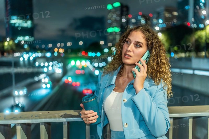 Professional businesswoman standing on a city bridge at night, having an important phone call with a reusable coffee cup in her hand, with urban traffic lights blurring in the background
