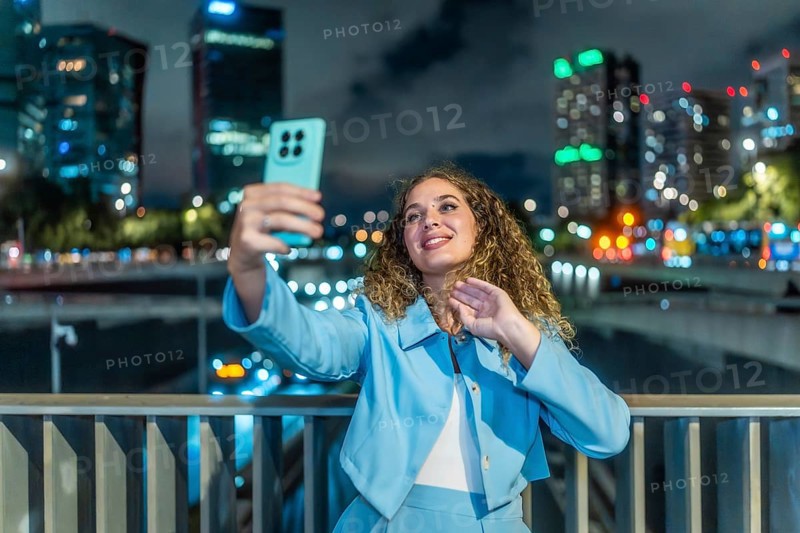 Young woman with curly hair and blue suit taking a selfie with her smartphone on a city bridge at night, bokeh lights from buildings and traffic visible in background