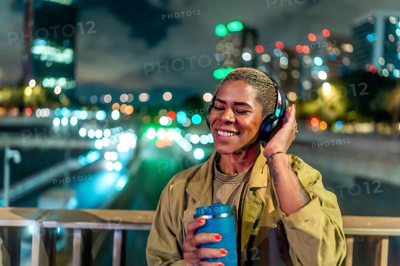 Woman standing on a bridge, holding a travel mug and wearing headphones while happily listening to music alone at night, with blurred city lights in the background