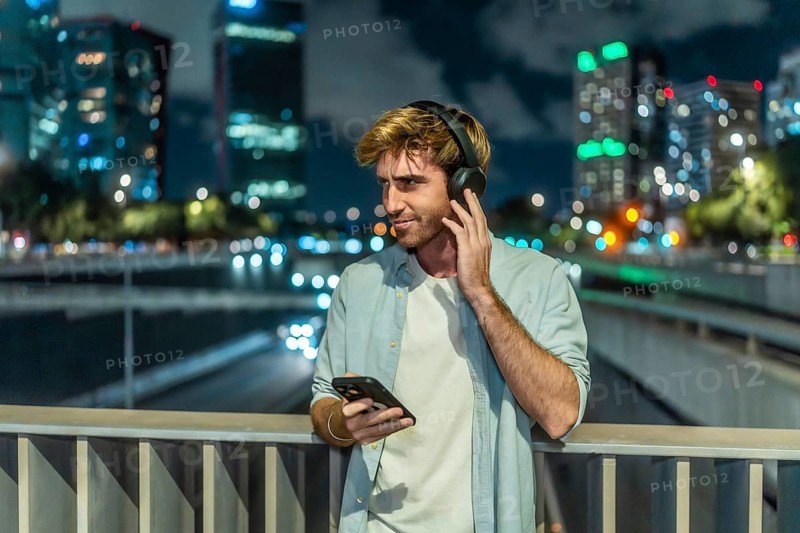 Young man is standing on a bridge, listening to music through headphones and holding a smartphone, with blurred city lights and traffic creating a vibrant bokeh background at night