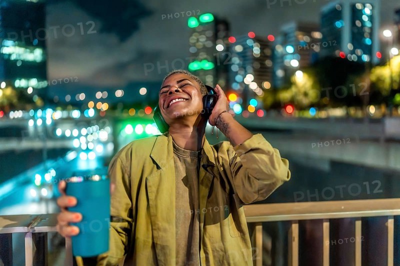 Young happy woman enjoying music through her headphones while standing on an urban bridge at night, with city lights and bokeh creating a vibrant background